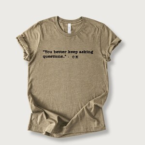 May include: A tan-colored t-shirt with the quote "You better keep asking questions..." - CK printed in black. The shirt has a crew neck and rolled-up sleeves, and is made of a soft, comfortable material.