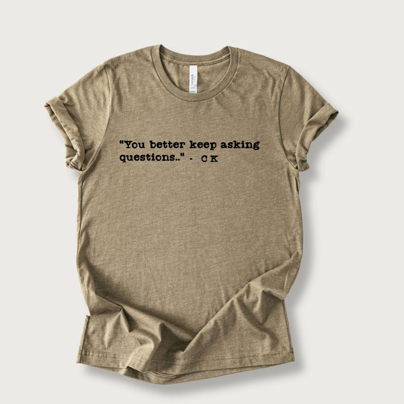 May include: A tan-colored t-shirt with the quote "You better keep asking questions..." - CK printed in black. The shirt has a crew neck and rolled-up sleeves, and is made of a soft, comfortable material.