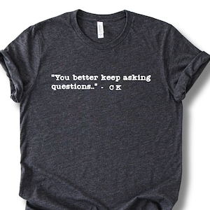 May include: Dark gray t-shirt with the quote "You better keep asking questions.." in white, followed by "- CK". The shirt has a crew neck and rolled-up sleeves. The fabric appears to be a soft, heathered material.