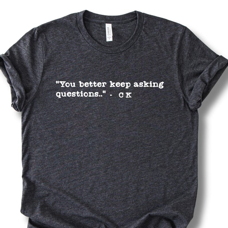May include: Dark gray t-shirt with the quote "You better keep asking questions.." in white, followed by "- CK". The shirt has a crew neck and rolled-up sleeves. The fabric appears to be a soft, heathered material.