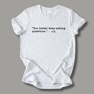 May include: Light gray t-shirt with the quote "You better keep asking questions.." - CK printed in black. The shirt is made of a soft material and has a crew neck.