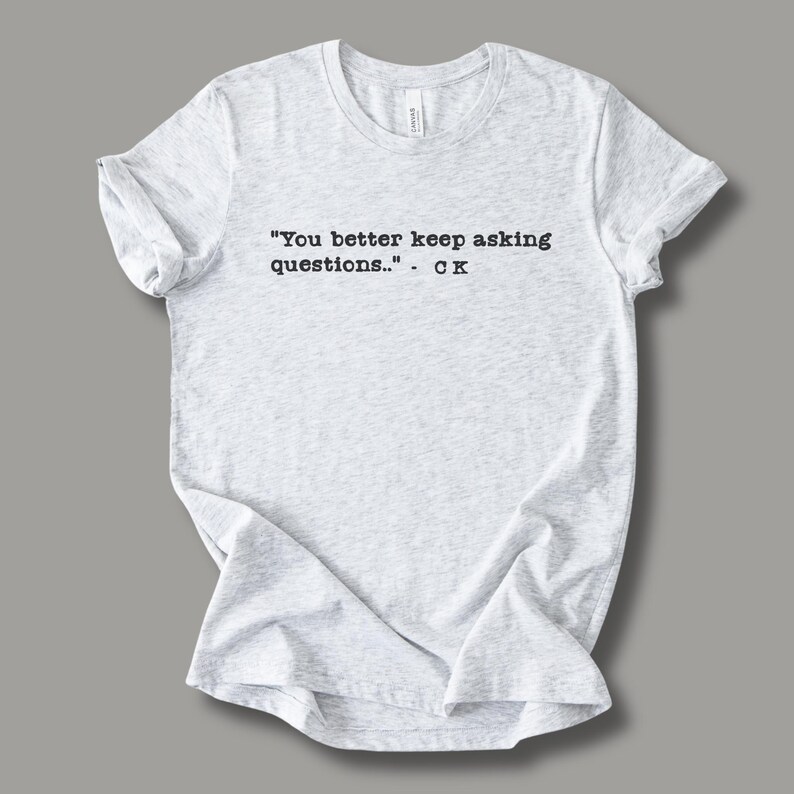 May include: Light gray t-shirt with the quote "You better keep asking questions.." - CK printed in black. The shirt is made of a soft material and has a crew neck.