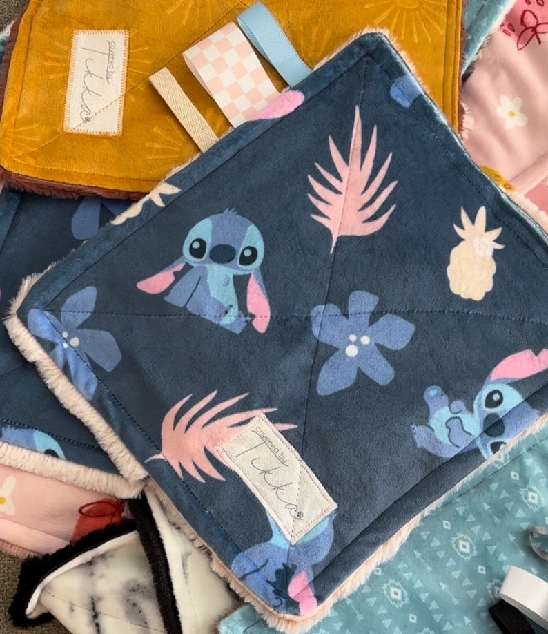 Puede incluir: Una colecci&oacute;n de mantas para beb&eacute;s con varios dise&ntilde;os. Una manta presenta un fondo azul oscuro con Stitch de Lilo & Stitch, hojas rosas y flores azules. Otras mantas son amarillas, rosas y estampadas. Cada manta tiene una etiqueta que dice "covered by Ikka".