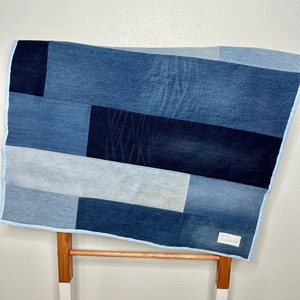Peut inclure: Une couverture en patchwork de denim avec des rectangles de denim bleu de différentes nuances. La couverture a une bordure bleu clair et une petite étiquette blanche avec du texte. La couverture est présentée sur un support en bois avec des pieds blancs.