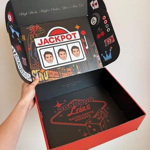 May include: A black and red cardboard gift box with a Las Vegas theme. The interior lid features a slot machine graphic with the text JACKPOT and three identical faces, plus casino icons like an 8-ball, cherries, and cards. The base reads WELCOME TO LEIA'S BACHELORETTE WEEKEND.