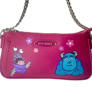 May include: A magenta-colored shoulder bag with a silver chain strap. The bag features illustrations of characters from the movie Monsters, Inc., including Boo and Sully, along with floral accents. The brand name "STEVE MADDEN" is displayed.