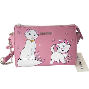 May include: Pink crossbody bag featuring illustrations of two white cats with pink bows and roses. The bag has a silver chain strap and the brand name "STEVE MADDEN" is visible.
