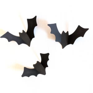 May include: Three black paper bats with folded wings are arranged against a white background. The bats are cut from a matte material and appear to be 3D. The bats are a Halloween decoration.
