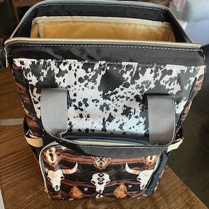 Pu&ograve; includere: Uno zaino nero e marrone con stampa in pelle di mucca e design a teschio e tribale. Lo zaino ha una chiusura a zip e due spalline regolabili.