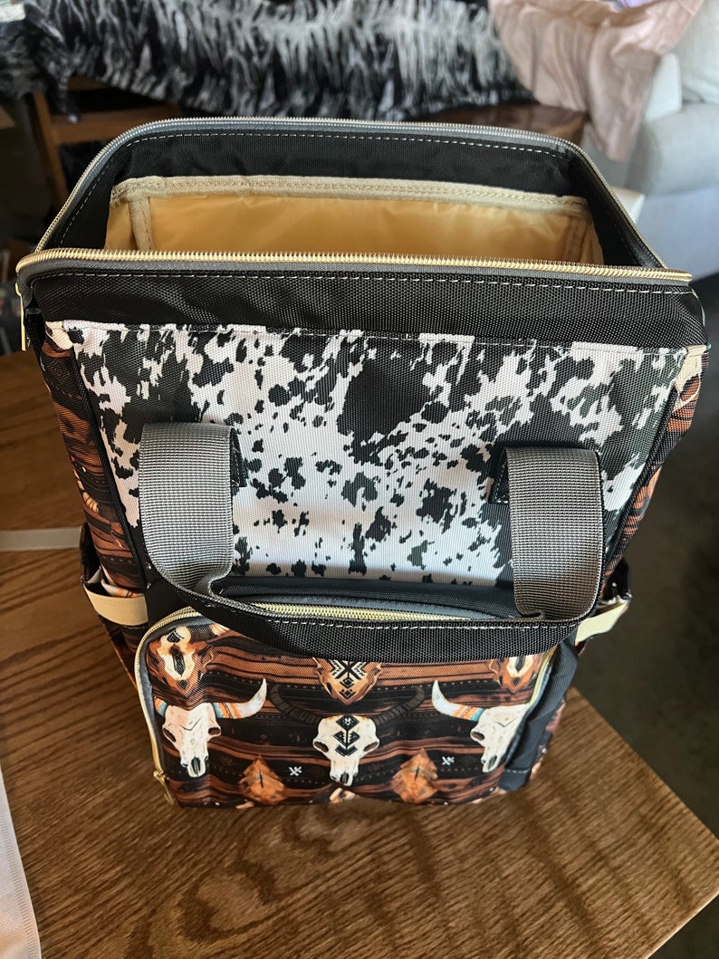 Pu&ograve; includere: Uno zaino nero e marrone con stampa in pelle di mucca e design a teschio e tribale. Lo zaino ha una chiusura a zip e due spalline regolabili.