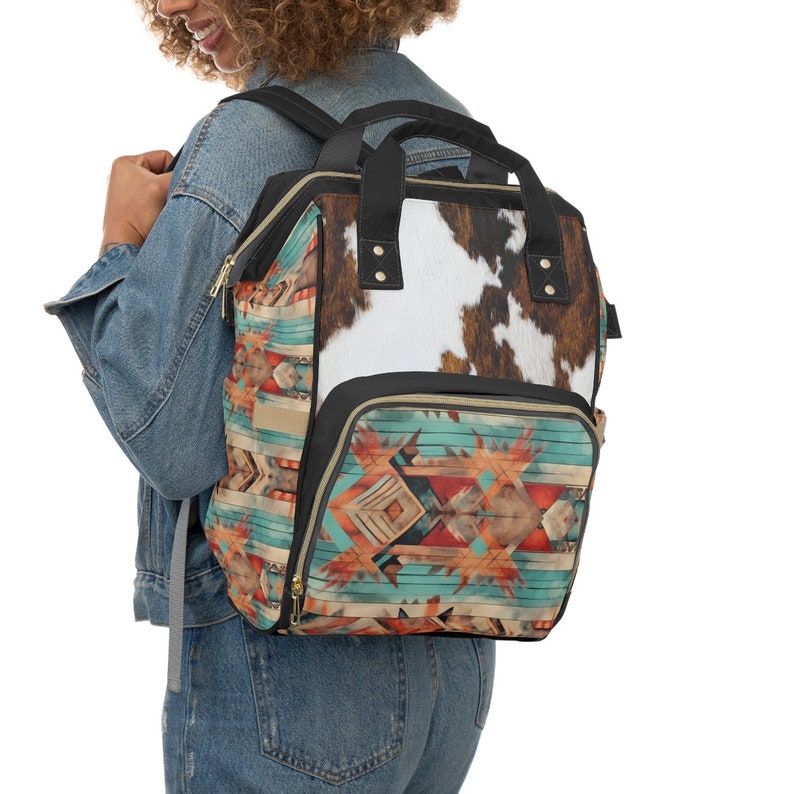 Pu&ograve; includere: Una borsa per pannolini con motivo nero e marrone con una patta in pelle di mucca. La borsa ha un ampio scomparto principale e diverse tasche pi&ugrave; piccole. Viene indossata da una persona che indossa una giacca di jeans e jeans.
