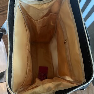 Pu&ograve; includere: L'interno di uno zaino nero con fodera beige. Lo zaino ha pi&ugrave; tasche e una chiusura a zip.