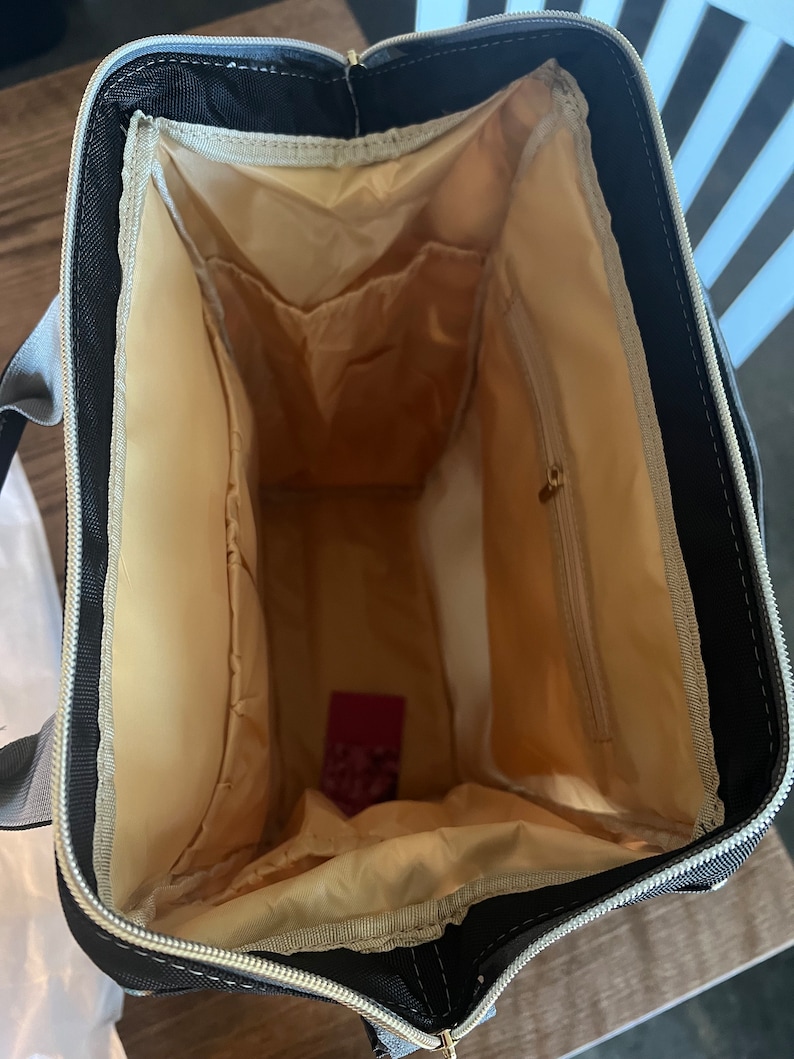 Pu&ograve; includere: L'interno di uno zaino nero con fodera beige. Lo zaino ha pi&ugrave; tasche e una chiusura a zip.