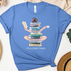 May include: A light blue t-shirt featuring a graphic of a person sitting on a stack of books, holding a book and a cup. The books have titles like "It Ends With Us" and "Layla." The text "IT'S A COHO THING" is at the bottom.