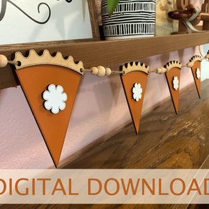 May include: A decorative garland featuring multiple orange triangular pie slice shapes with scalloped crust edges and white flower-shaped accents, strung together with natural twine and interspersed with round wooden beads.