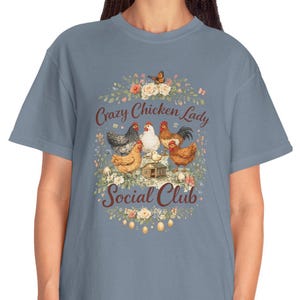 以下が含まれることがあります： 鶏のグラフィックと「Crazy Chicken Lady Social Club」の文字が花で囲まれた、青みがかったグレーのTシャツ。カジュアルなスタイルで、普段使いに適しています。