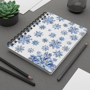 May include: A white spiral bound notebook with a blue watercolor snowflake pattern. The notebook is lying on a gray surface with a pencil and a succulent plant in a black pot.