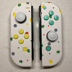 Puede incluir: Dos mandos Joy-Con blancos para Nintendo Switch. Los mandos están decorados con iconos y patrones coloridos, incluyendo hojas verdes, casas amarillas y puntos rojos. Los botones son amarillos y verdes.
