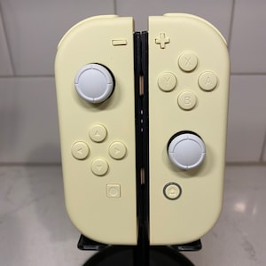 May include: A yellow Nintendo Switch Joy-Con controller with white buttons. The controller is on a black stand.