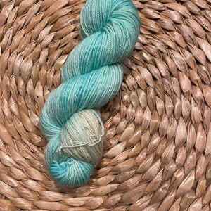 May include: A skein of light blue and white yarn with a gradient effect. The yarn is wound into a loose coil and is sitting on a woven basket.