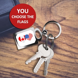May include: A silver keychain with a heart-shaped charm featuring the Canadian and American flags. The keychain is attached to a set of keys. The text "YOU CHOOSE THE FLAGS" is visible in a red circle.