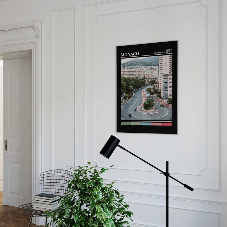 Peut inclure: Une affiche noir et blanc avec le mot "MONACO" en blanc en haut. L'affiche pr&eacute;sente une photographie d'une route sinueuse avec des b&acirc;timents et des arbres en arri&egrave;re-plan. Le texte "EUROPE" et "43.7384&deg; N, 7.4208&deg; E" sont en blanc plus petit en haut de l'affiche.