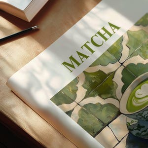 May include: A close-up of a magazine cover with the word "MATCHA" printed in green letters. The background is a green and white tiled pattern with a cup of green tea with latte art on top of the tiles.