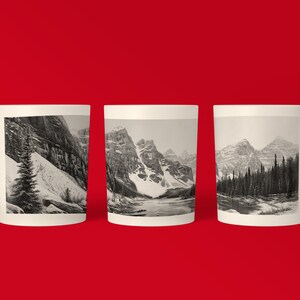 May include: Three white ceramic mugs with black and white images of snow-capped mountains and a lake. The mugs are arranged in a row on a red background.
