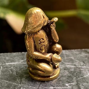 Könnte beinhalten: Kleine, messingfarbene Figur eines sitzenden Affen. Der Affe sitzt in einer Hockstellung, mit den Händen am Gesicht und einem kleinen, kugelartigen Objekt. Die Figur hat geätzte Details und eine glatte, polierte Oberfläche. Der Kopf des Affen ist leicht geneigt.