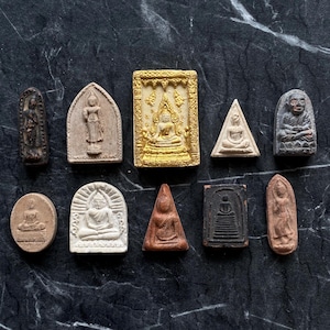 Può includere: Una collezione di piccoli amuleti di Buddha, finemente dettagliati, in varie forme e colori. Gli amuleti sono realizzati con materiali come argilla e pietra, alcuni con accenti dorati. La collezione comprende forme rettangolari, ovali e triangolari.