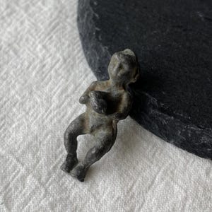 Könnte beinhalten: Kleine Metallfigur im antiken Stil, die eine sitzende Figur darstellt, möglicherweise ein Cherub oder Putto. Die Figur hat eine gedämpfte grau-grüne Farbe mit sichtbaren Details von Gesicht, Armen und Beinen. Die Figur hält einen Gegenstand in ihren Armen.