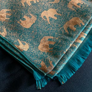 May include: Teal and gold decorative fabric with an elephant pattern. The fabric is folded, revealing a fringed edge. The elephants are gold and the background is teal.
