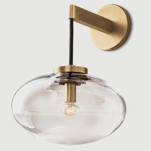 May include: A modern wall sconce featuring a clear, oval-shaped glass shade and a brass-toned metal frame. The fixture includes a brass-colored bulb holder and a black cord. The sconce is mounted on a round brass-colored base.