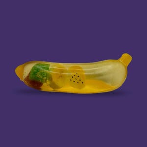 Può includere: Un oggetto traslucido a forma di banana realizzato in resina o vetro. L'interno contiene elementi decorativi incorporati tra cui un quadrato verde, una fetta circolare gialla e una sezione gialla con piccoli punti neri.