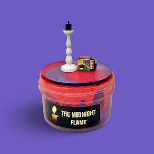 May include: A clear plastic container filled with red and purple slime, topped with a white miniature candlestick and a small matchbox. A black label on the front features a yellow candle illustration and the text THE MIDNIGHT FLAME in white.