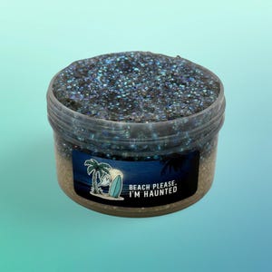 May include: A clear plastic jar filled with dark blue and black textured slime containing small beads. A rectangular label on the front features a palm tree, a surfboard, a ghost, and the text "BEACH PLEASE, I'M HAUNTED" in white letters.