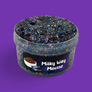 May include: A clear plastic container filled with black slime, speckled with iridescent glitter. The container has a label that reads "Milky Way Mousse" with a graphic of a bowl of chocolate mousse. The background is a solid purple.