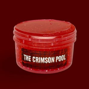 Può includere: Un contenitore rotondo in plastica trasparente riempito con una sostanza gelatinosa rosso brillante e glitterata. Un'etichetta rettangolare sulla parte anteriore presenta un motivo a griglia nera e il testo bianco THE CRIMSON POOL in carattere colante.