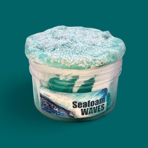 Può includere: Un contenitore di plastica trasparente pieno di slime blu e bianco. Lo slime ha una superficie strutturata con piccole perle bianche. Il contenitore ha un'etichetta con la scritta "Seafoam WAVES" con una grafica a onde.