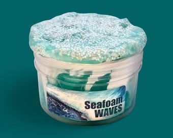 May include: A clear plastic pot filled with blue and white slime. The slime has a textured surface with small white beads. The pot has a label that reads "Seafoam WAVES" with a wave graphic.