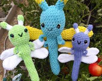 May include: Three crocheted dragonfly toys. The dragonfly on the left is green with white wings. The dragonfly in the center is blue with yellow wings. The dragonfly on the right is purple with white wings.