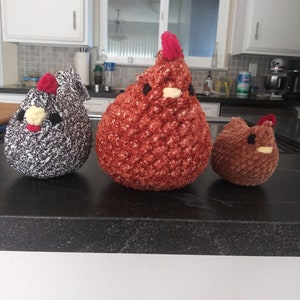 May include: Three crocheted chicken plushies. One is brown, one is gray with white speckles, and one is red with a white beak and yellow eyes.