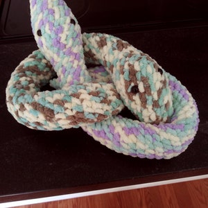 May include: Two crocheted snakes, one in brown, white, and green, and the other in purple, white, and green, are intertwined on a black surface.