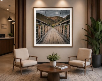 Könnte beinhalten: Ein quadratischer Kunstdruck in einem Rahmen zeigt eine hölzerne Hängebrücke, die unter einem bewölkten Himmel auf hügelige Landschaften zuführt. Das Bild hängt in einem Wartebereich mit zwei beigen Sesseln, einem runden Holztisch und einer Pflanze.