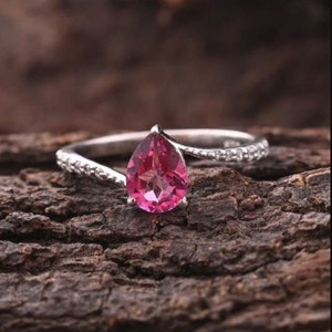 Könnte beinhalten: Ein silberner Ring mit einem birnenförmigen, rosa Edelstein. Das Band ist mit kleinen, klaren Steinen verziert. Der Ring ist vor einem strukturierten, braunen Hintergrund platziert, der das Design und die Farbe hervorhebt.