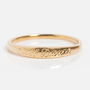 Könnte beinhalten: Ein goldfarbener Ring mit der Gravur "Till Death". Der Ring ist ein schlichtes Banddesign, wobei der Text um die Außenseite des Rings verläuft. Der Ring ist vor einem weißen Hintergrund abgebildet.