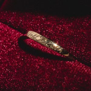 Könnte beinhalten: Ein goldener Ring mit der Inschrift "Till Death" in einer eleganten Schrift. Der Ring ist vor einem tiefroten, glitzernden Hintergrund platziert, was einen auffälligen Kontrast erzeugt. Das Design des Rings ist schlicht, aber markant.