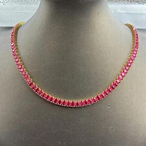 Op de afbeelding: Een goudkleurige ketting met een rij ovaal geslepen, helderrode edelstenen. De edelstenen zijn dicht op elkaar geplaatst, waardoor een doorlopende lijn van kleur ontstaat. De ketting wordt gepresenteerd op een neutrale achtergrond.