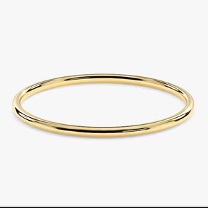 Könnte beinhalten: Ein einfacher, eleganter Goldarmreif. Das runde, polierte Armband ist ein massives Goldband, das das Licht reflektiert. Der Schmuck ist vor einem schlichten weißen Hintergrund platziert, der sein minimalistisches Design hervorhebt.
