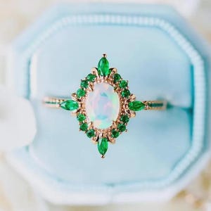 Könnte beinhalten: Ein goldener Ring mit einem ovalen, schillernden Opal-Mittelstein, umgeben von kleinen, grünen Smaragden. Der Ring hat ein florales Design mit Smaragd-Akzenten, vor hellblauem Hintergrund.
